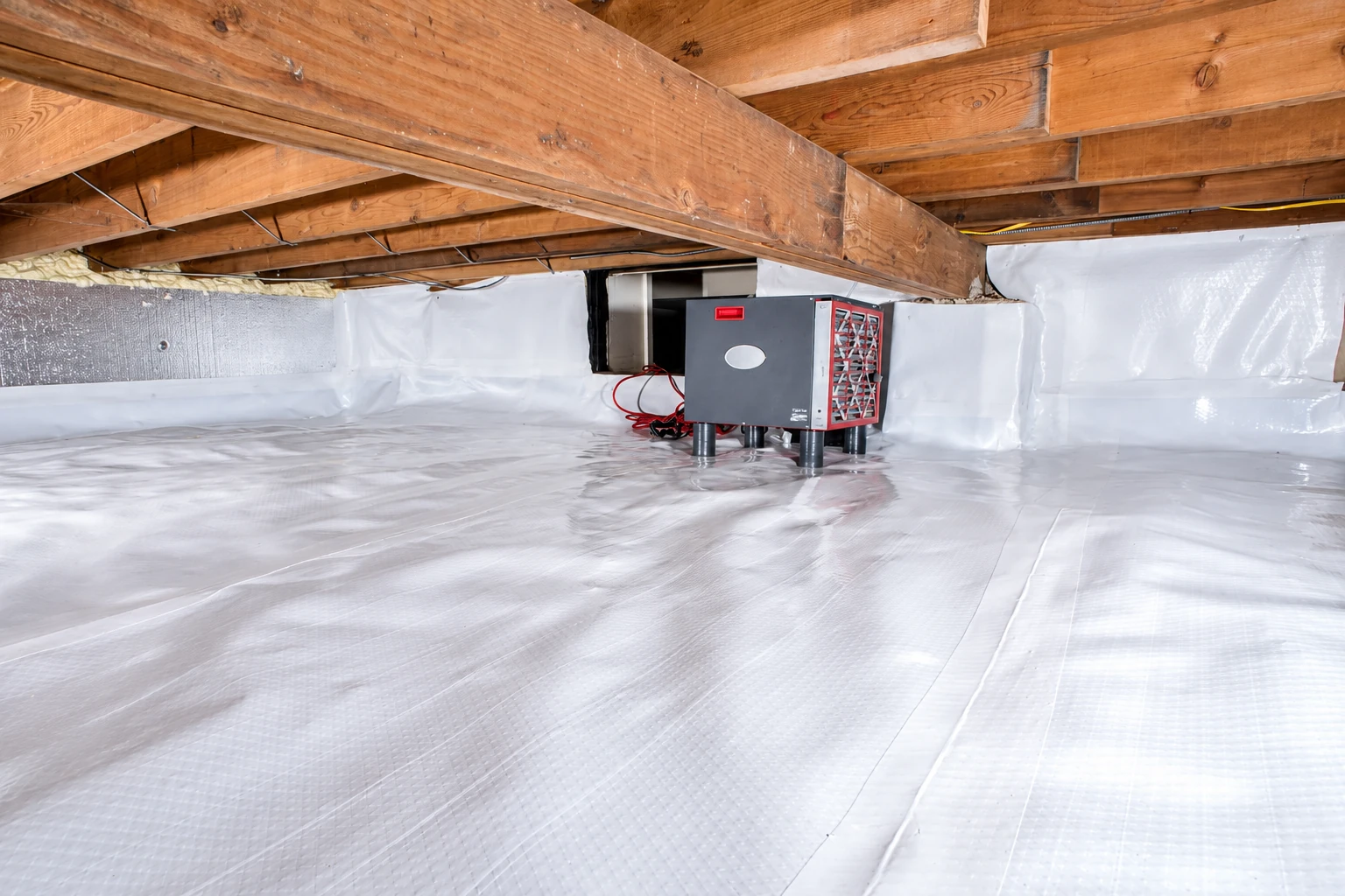 A compact machine sits on white vapor barrier sheeting in a crawl space enclosed by exposed wooden beams.