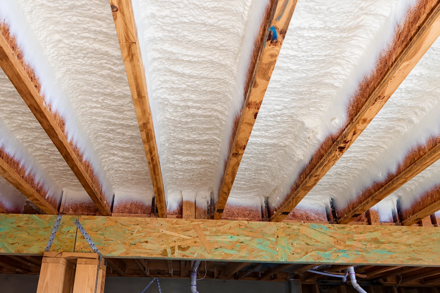 White spray foam fills the spaces between exposed wooden roof rafters above an unfinished ceiling.