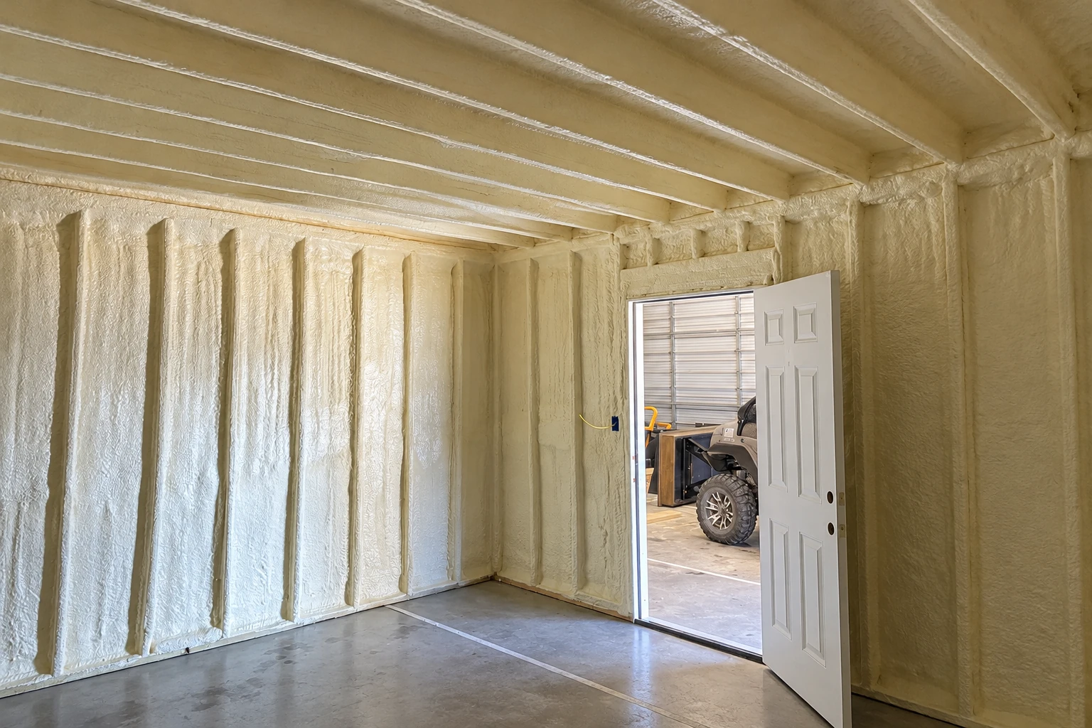 Cream-colored spray foam coats the walls and ceiling of a garage with an open door.