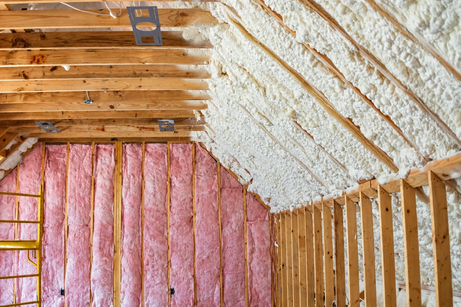 White spray foam covers a sloped attic roof beside exposed rafters and pink batt insulation in the walls.