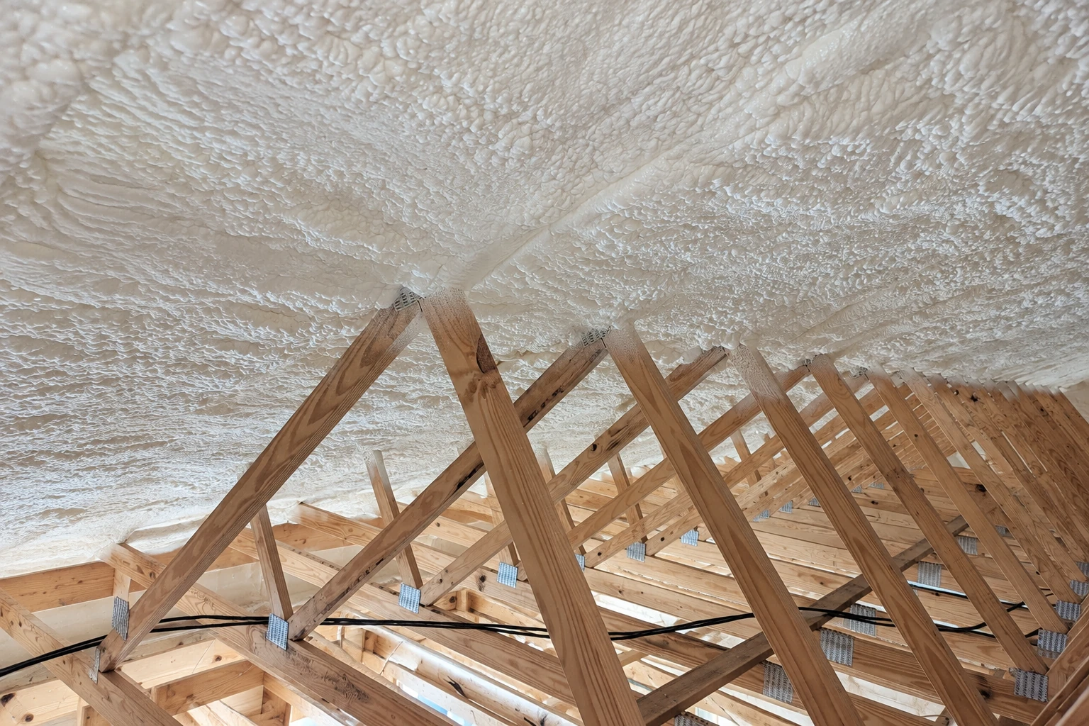 White spray foam coats the underside of a roof above a large framework of exposed wooden trusses.