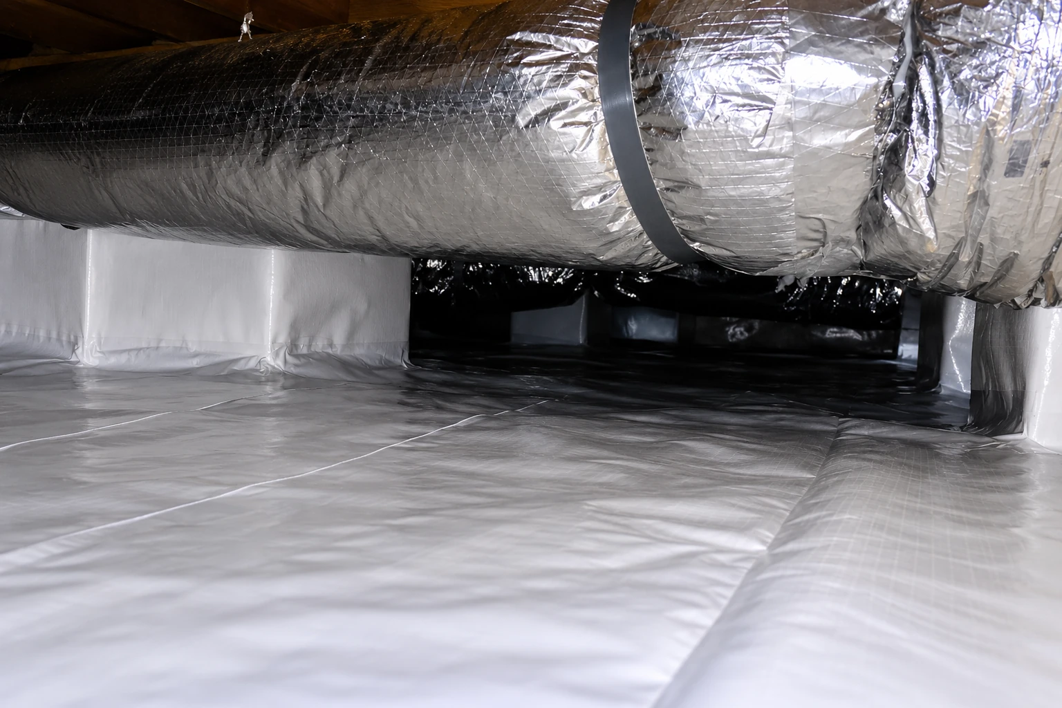 Vapor barrier installation in a crawl space, with reflective sheeting covering the floor and ductwork overhead.